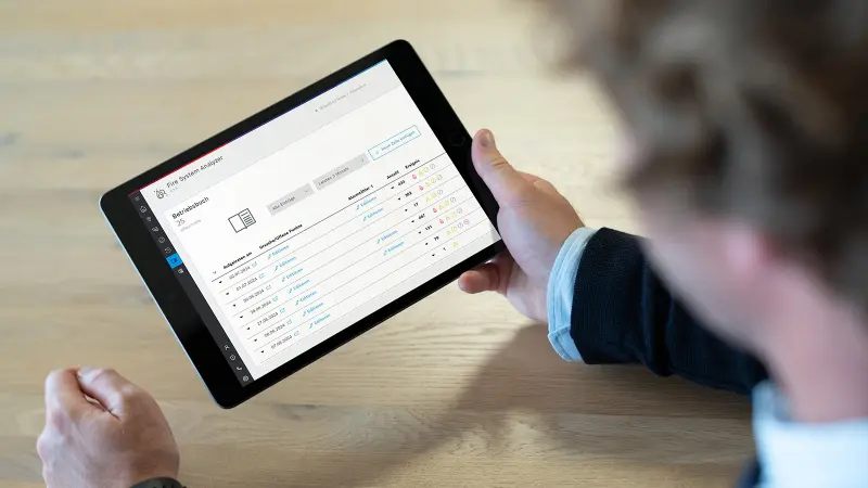 Person hält Tablet mit Fire System Analyzer-Anwendung, die ein Betriebsbuch mit chronologisch aufgelisteten Ereignissen und Alarmzählern vom Juni und Juli 2024 anzeigt. Die Tabelle enthält Datumsangaben, Statuswerte und verschiedene Ereignissymbole.