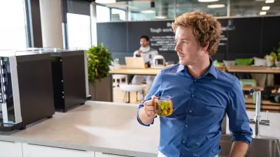 Man with curly reddish-blond hair in a blue shirt holding a glass of tea, standing in a modern office kitchen with Datanaut in the background.