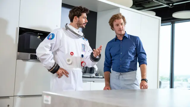 NEXOSPACE Datanaut holds a black tablet and points to it; on the right stands a man with curly brown hair in a blue shirt, gesturing in a modern office environment.