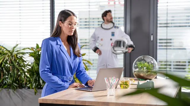 Frau in blauem Anzug arbeitet am Laptop im Büro während Astronaut im Hintergrund steht.