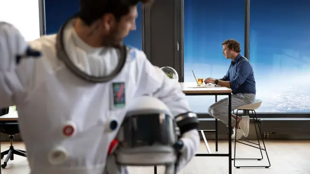 Ein Gebäudemanager sitzt am Schreibtisch und arbeitet an seinem Laptop. Im Vordergrund ist ein Datanaut mit Helm zu sehen – als Visualisierung für Sicherheit durch digitalen Brandschutz.