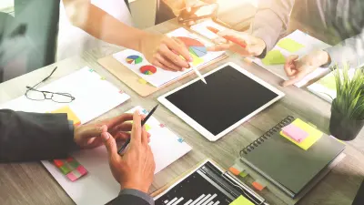 Blick auf einen Schreibtisch von oben, an dem drei Personen stehen, mit Blättern mit Diagrammen, Post-its und Blöcken.