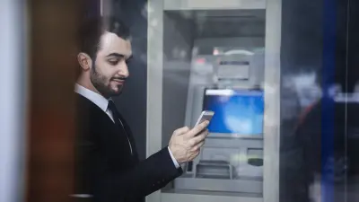 Dunkelhaariger Mann mit Bart steht vor einem Geldautomaten mit Smartphone in der Hand.