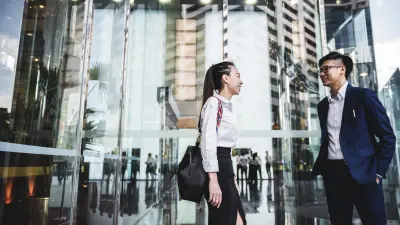 Mann und Frau in Business Kleidung stehen von einem Eingang eines gläsernen Gebäudes.
