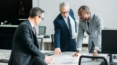 Drei Männer mit Anzug und Brille stehen um einen Tisch, auf dem ein Plan liegt.