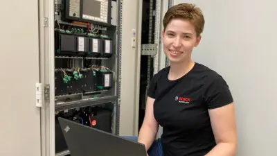 Sabine schaut in die Kamera, hat ihren PC auf dem Schoß und neben ihr ist der Schaltschrank zu sehen