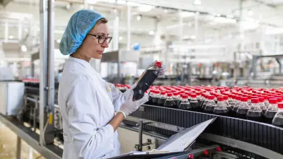 Women with smock and hood looking at a filled bottle that she has taken from a conveyor belt full of other filled bottles