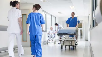 Alcuni operatori che camminano lungo il corridoio di un ospedale, uno trasportando una barella e l'altro aiutando una donna su una sedia a rotelle.
